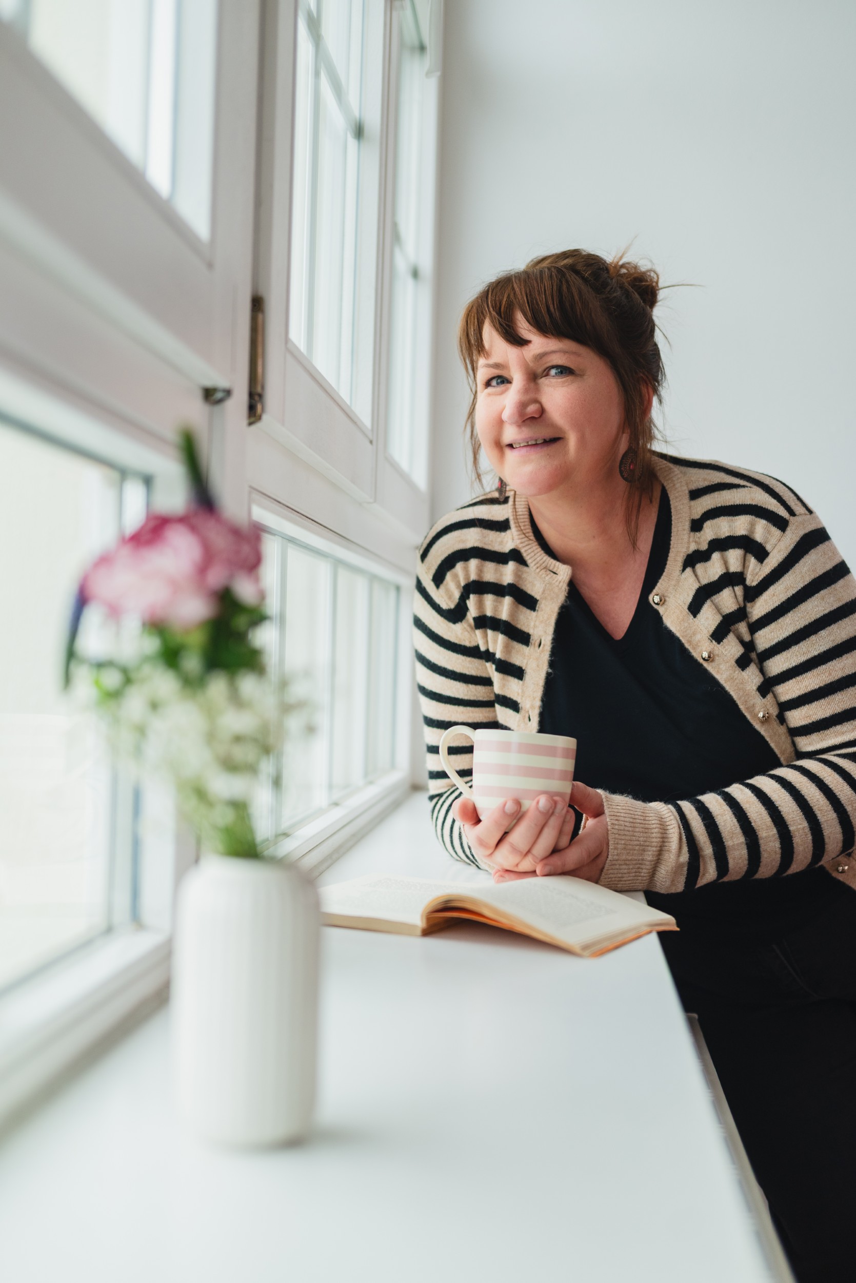 Anja Zwade am Fenster mit Buch und Tasse, daneben eine Vase mit Blumen.