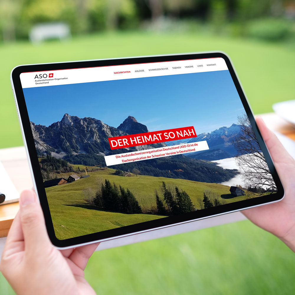 Auf einem Tablet: Schweizer Bergpanorama mit Almwiese und dem Titel „Der Heimat so nah“.